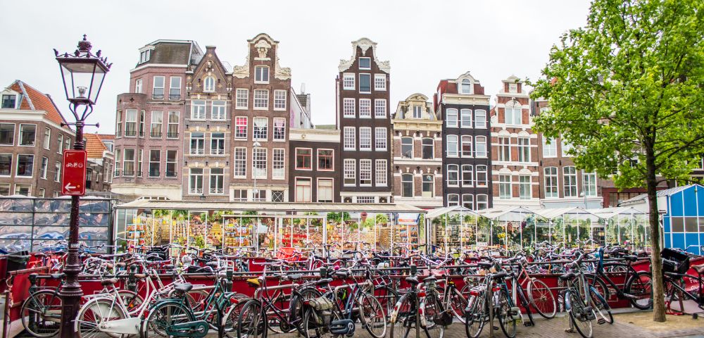 A large outdoor bicycle parking lot is in the foreground, with numerous bikes lined up. Behind the bikes are several glass-enclosed market stalls filled with goods, and beyond that are tall, narrow historic buildings with distinct gabled roofs under a cloudy sky.