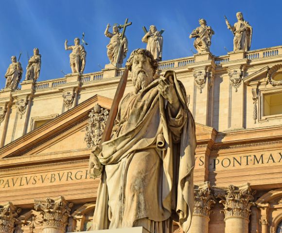 A weathered stone statue of a robed man holding a sword stands in the foreground, with the facade of a grand building featuring statues along its upper balustrade in the background.