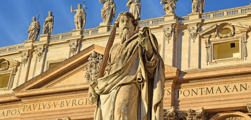 A weathered stone statue of a robed man holding a sword stands in the foreground, with the facade of a grand building featuring statues along its upper balustrade in the background.