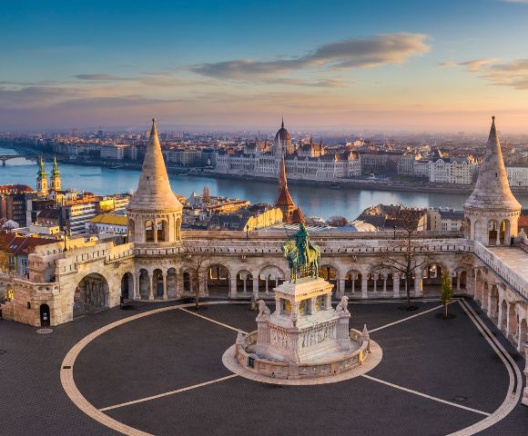 A panoramic sunrise view of Fisherman's Bastion in Budapest, Hungary, with its fairytale towers and a statue in the foreground, overlooking the Danube River and the Parliament Building.