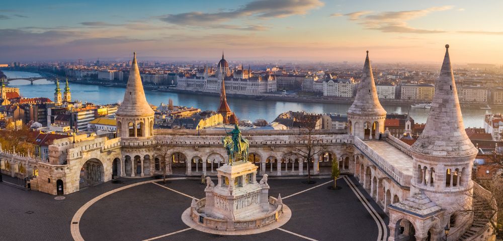 A panoramic sunrise view of Fisherman's Bastion in Budapest, Hungary, with its fairytale towers and a statue in the foreground, overlooking the Danube River and the Parliament Building.