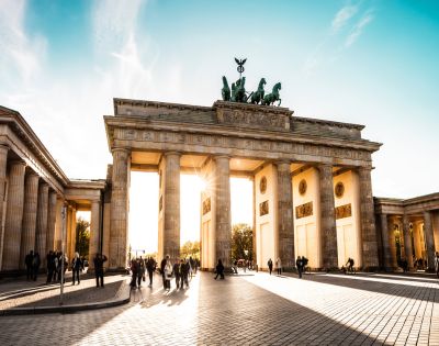 The Brandenburg Gate in Berlin, Germany, with sunburst light through its central arch and people walking in the plaza below.