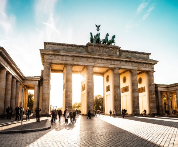 The Brandenburg Gate in Berlin, Germany, with sunburst light through its central arch and people walking in the plaza below.