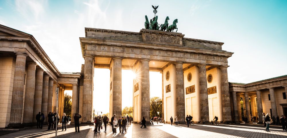 The Brandenburg Gate in Berlin, Germany, with sunburst light through its central arch and people walking in the plaza below.