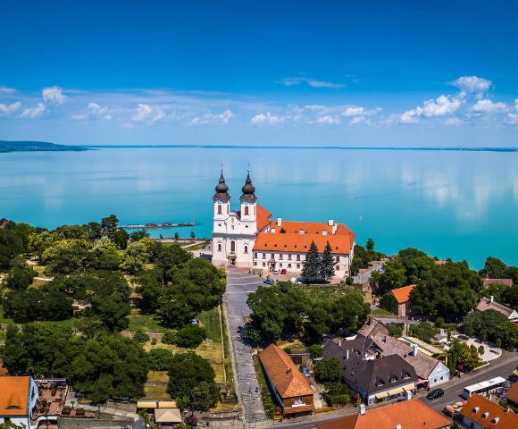 Aerial view of a white Benedictine abbey with twin onion domes and an orange tiled roof, situated on a hill overlooking a vast, turquoise lake under a bright blue sky with scattered white clouds.