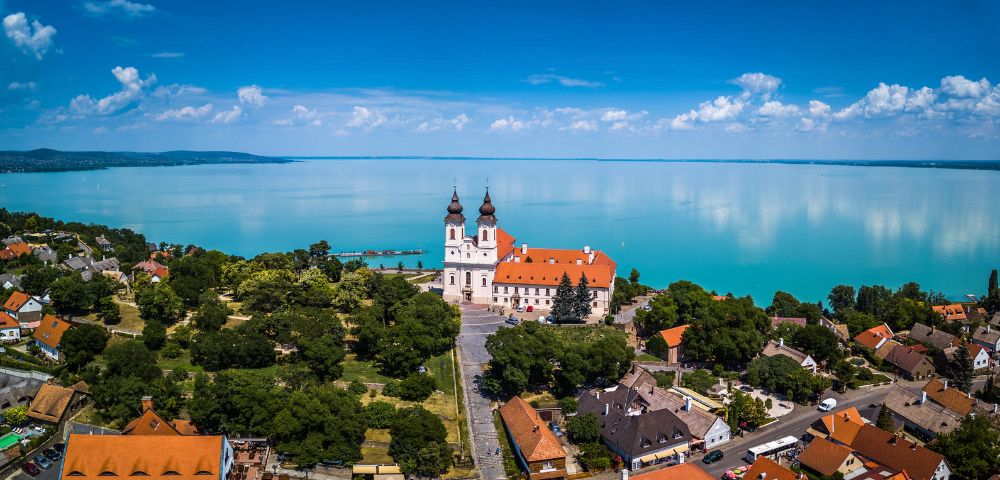 Aerial view of a white Benedictine abbey with twin onion domes and an orange tiled roof, situated on a hill overlooking a vast, turquoise lake under a bright blue sky with scattered white clouds.