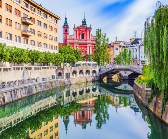 A picturesque canal with the reflection of a pink baroque church and surrounding buildings on a bright, sunny day.