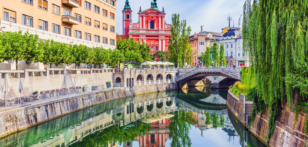 A picturesque canal with the reflection of a pink baroque church and surrounding buildings on a bright, sunny day.