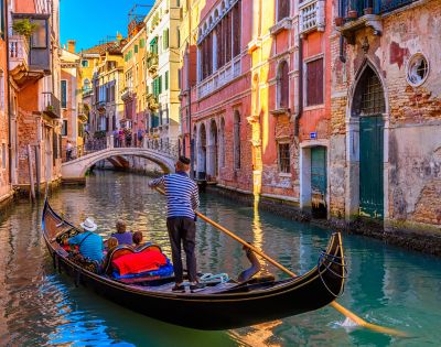A gondola glides through a narrow canal in Venice, Italy, with a gondolier in a striped shirt steering with a long oar. Ornate, colorful buildings line the waterway, and a pedestrian bridge arches over the canal in the background.