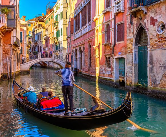 A gondola glides through a narrow canal in Venice, Italy, with a gondolier in a striped shirt steering with a long oar. Ornate, colorful buildings line the waterway, and a pedestrian bridge arches over the canal in the background.