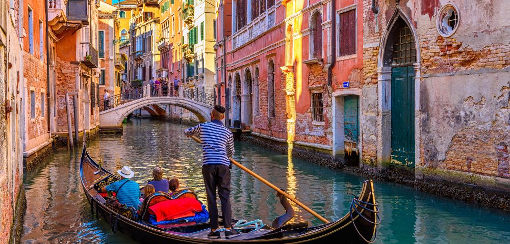 A gondola glides through a narrow canal in Venice, Italy, with a gondolier in a striped shirt steering with a long oar. Ornate, colorful buildings line the waterway, and a pedestrian bridge arches over the canal in the background.