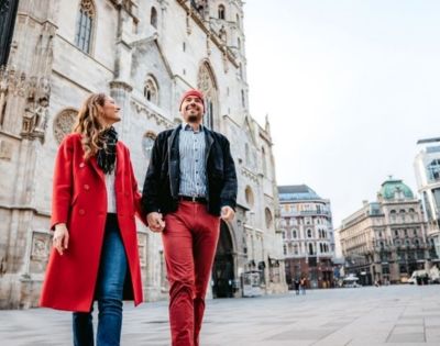 A smiling couple holding hands walks forward on a paved city square in front of a grand Gothic-style cathedral and other historic buildings.