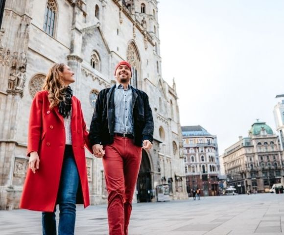 A smiling couple holding hands walks forward on a paved city square in front of a grand Gothic-style cathedral and other historic buildings.
