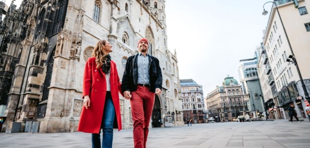 A smiling couple holding hands walks forward on a paved city square in front of a grand Gothic-style cathedral and other historic buildings.