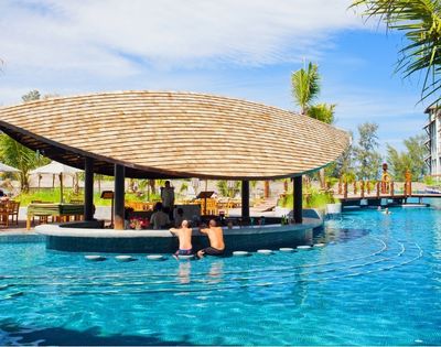 A swim-up bar with a thatched roof is set in a bright blue swimming pool at a tropical resort, with people sitting at the bar and swimming.