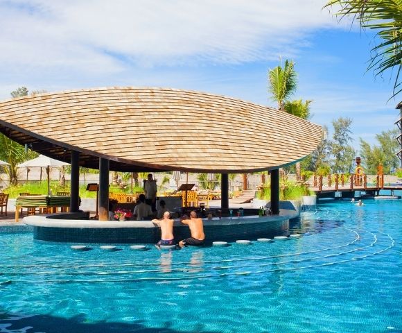 A swim-up bar with a thatched roof is set in a bright blue swimming pool at a tropical resort, with people sitting at the bar and swimming.