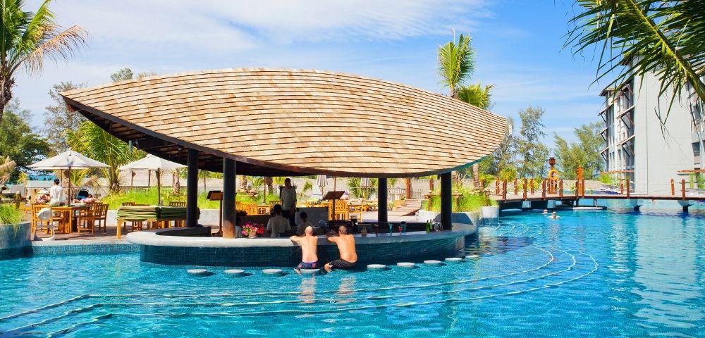 A swim-up bar with a thatched roof is set in a bright blue swimming pool at a tropical resort, with people sitting at the bar and swimming.