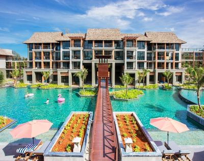A modern resort hotel with multiple buildings, a large swimming pool with island landscaping, a wooden walkway, and inflatable pool toys.