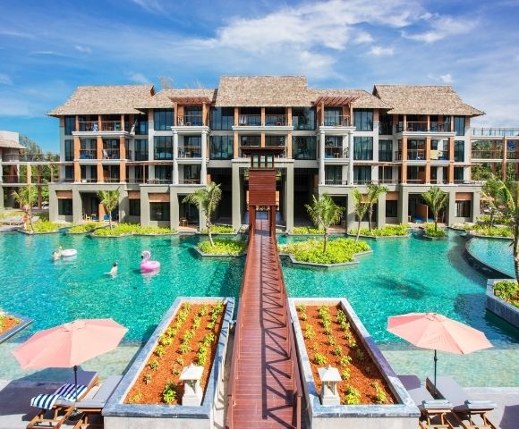 A modern resort hotel with multiple buildings, a large swimming pool with island landscaping, a wooden walkway, and inflatable pool toys.