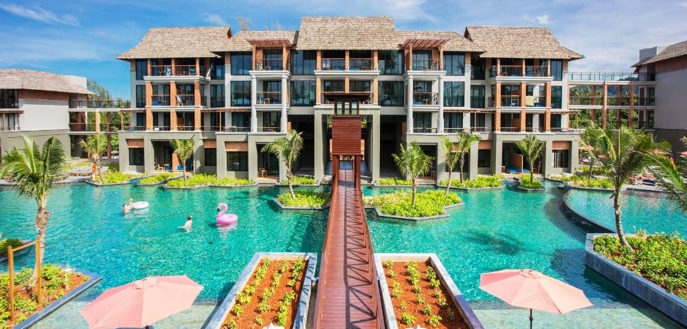A modern resort hotel with multiple buildings, a large swimming pool with island landscaping, a wooden walkway, and inflatable pool toys.