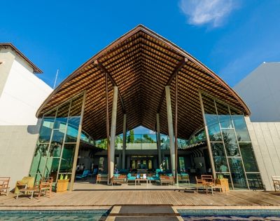 Modern open-air resort lobby with a large thatched roof, glass walls, and comfortable seating around a pool.