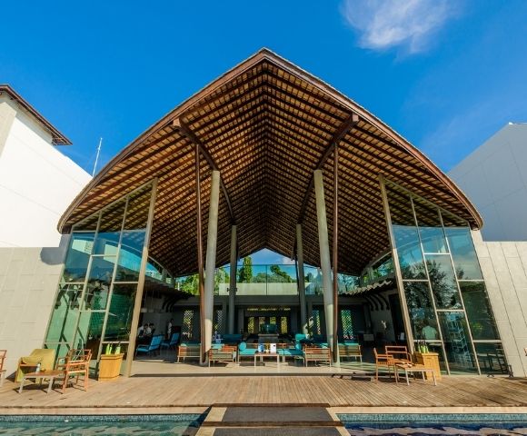 Modern open-air resort lobby with a large thatched roof, glass walls, and comfortable seating around a pool.