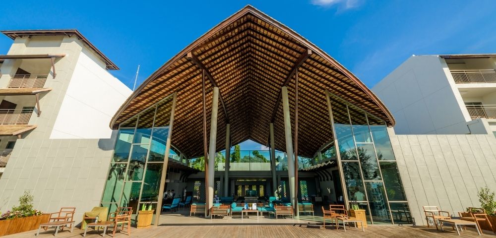 Modern open-air resort lobby with a large thatched roof, glass walls, and comfortable seating around a pool.