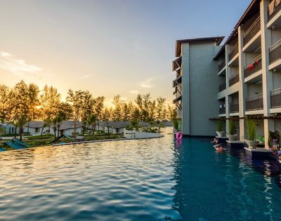 Infinity pool beside a modern hotel building at sunset with trees and small houses in the background.
