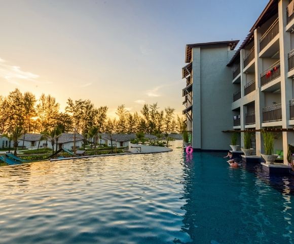 Infinity pool beside a modern hotel building at sunset with trees and small houses in the background.