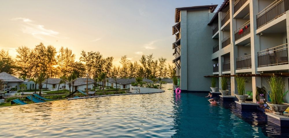 Infinity pool beside a modern hotel building at sunset with trees and small houses in the background.