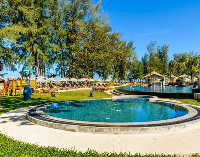 Outdoor resort pool area with circular pools, lounge chairs with umbrellas, playground, and trees under a clear blue sky.
