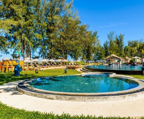 Outdoor resort pool area with circular pools, lounge chairs with umbrellas, playground, and trees under a clear blue sky.
