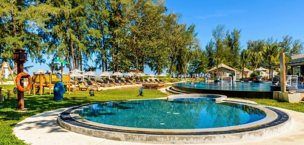 Outdoor resort pool area with circular pools, lounge chairs with umbrellas, playground, and trees under a clear blue sky.