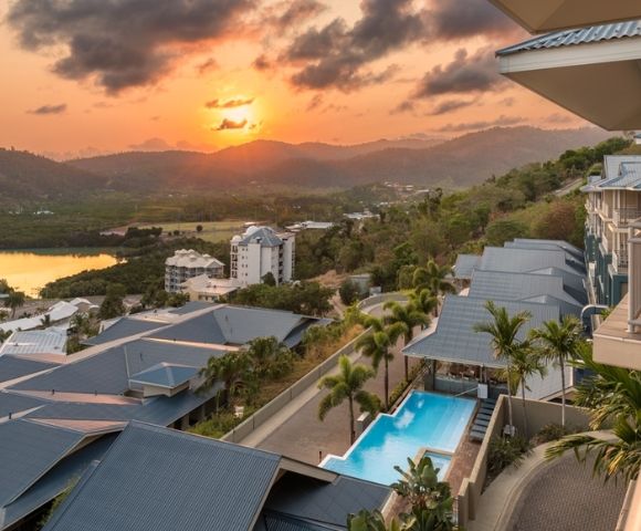 Resort buildings and a swimming pool with a tropical landscape and sunset over mountains and water.