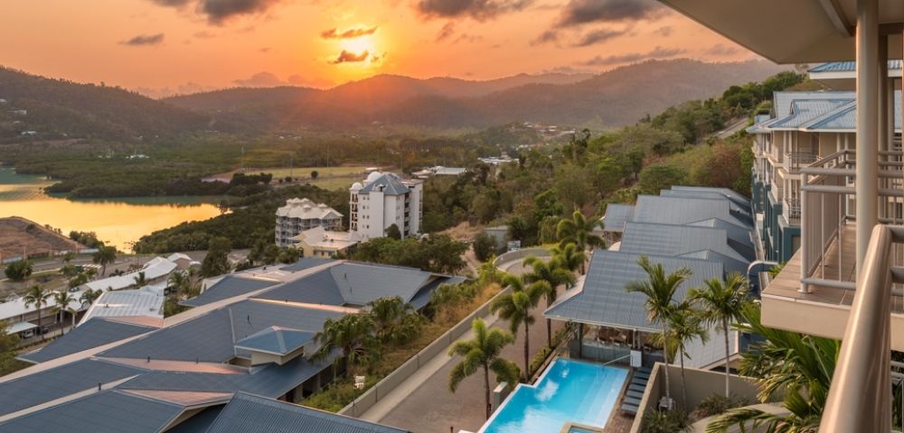Resort buildings and a swimming pool with a tropical landscape and sunset over mountains and water.