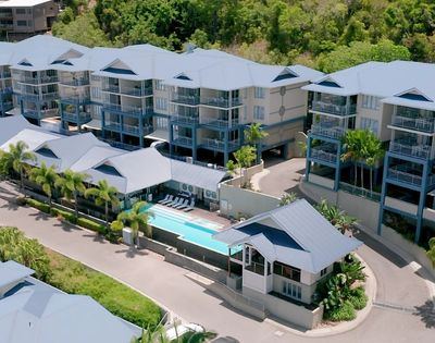 Aerial view of a resort with multiple buildings featuring blue balconies and light-colored roofs, a swimming pool, lounge chairs, and surrounding lush green foliage.