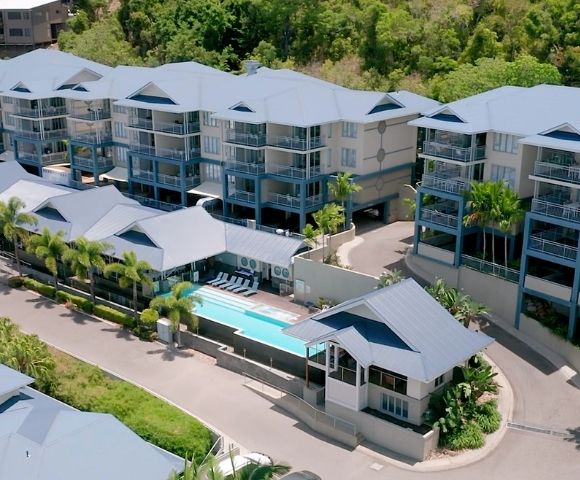 Aerial view of a resort with multiple buildings featuring blue balconies and light-colored roofs, a swimming pool, lounge chairs, and surrounding lush green foliage.