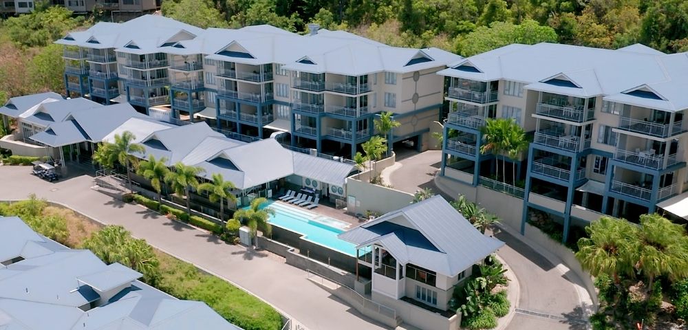 Aerial view of a resort with multiple buildings featuring blue balconies and light-colored roofs, a swimming pool, lounge chairs, and surrounding lush green foliage.