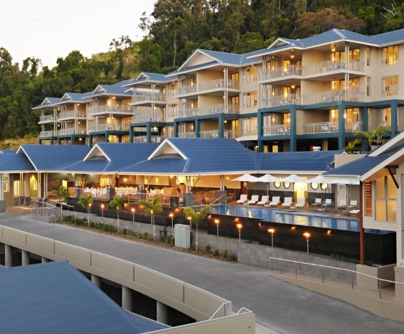 A modern resort complex with multiple levels of rooms and balconies, featuring blue roofs and a swimming pool area with lounge chairs.
