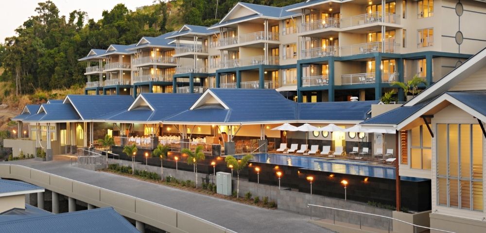 A modern resort complex with multiple levels of rooms and balconies, featuring blue roofs and a swimming pool area with lounge chairs.