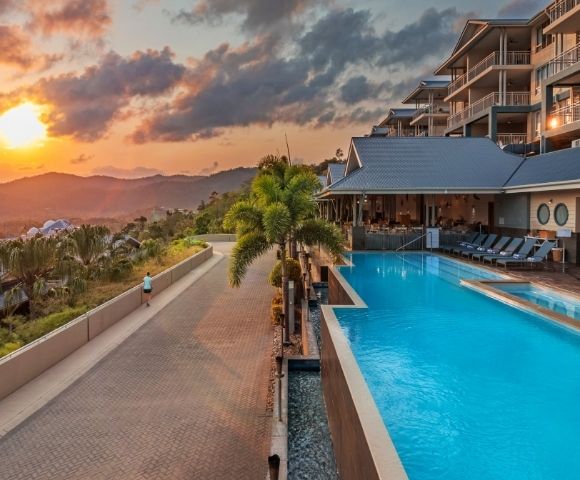 Resort pool with turquoise water, lounge chairs, and palm trees at sunset with mountains in the background.