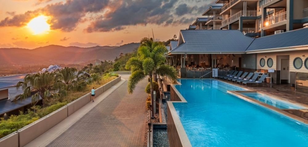 Resort pool with turquoise water, lounge chairs, and palm trees at sunset with mountains in the background.