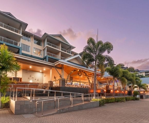 A multi-story resort building with balconies and palm trees, illuminated by warm lighting in the evening.