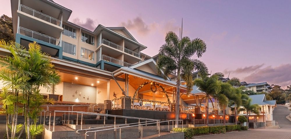 A multi-story resort building with balconies and palm trees, illuminated by warm lighting in the evening.