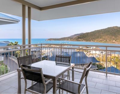 Balcony with a table and chairs overlooking a bay with sailboats and a hilly coastline.