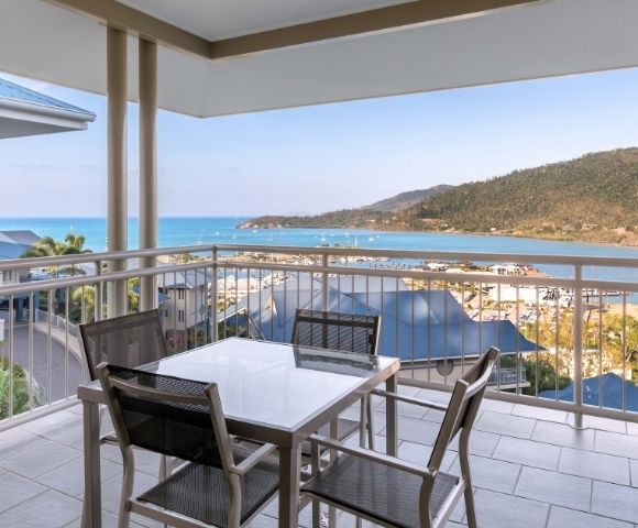 Balcony with a table and chairs overlooking a bay with sailboats and a hilly coastline.