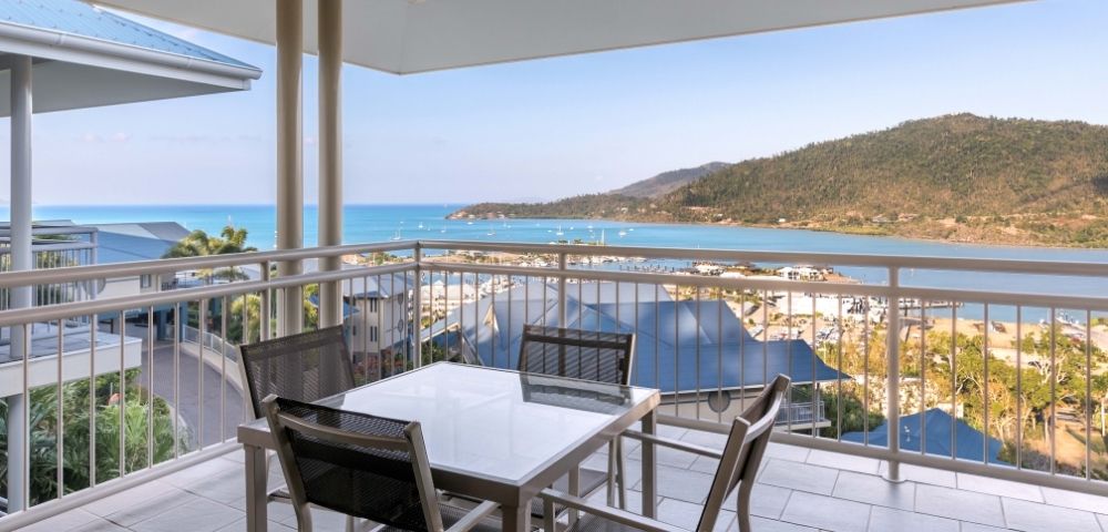 Balcony with a table and chairs overlooking a bay with sailboats and a hilly coastline.