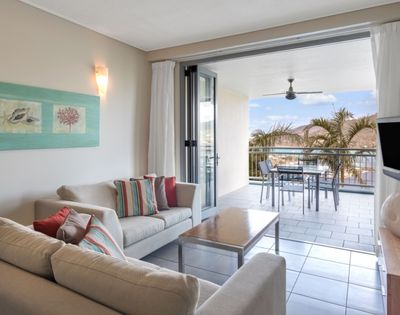 Living room with a sofa, coffee table, and a large sliding glass door opening to a balcony with a table and chairs, overlooking palm trees and a coastal view.
