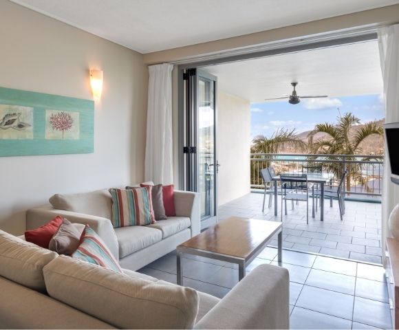 Living room with a sofa, coffee table, and a large sliding glass door opening to a balcony with a table and chairs, overlooking palm trees and a coastal view.