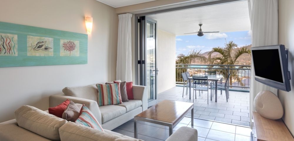 Living room with a sofa, coffee table, and a large sliding glass door opening to a balcony with a table and chairs, overlooking palm trees and a coastal view.